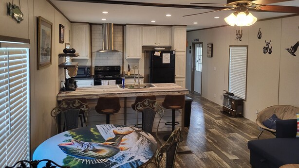 A modern kitchen with white cabinets, a black refrigerator, and a dining area with a round table featuring a fish decal. - Cozy 3-bedroom 2 bath,5beds mobile home. Close to a boat launch. Covered parking (Many)
