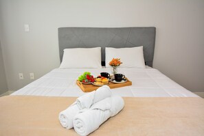 Una cama bien hecha con edredón blanco, una bandeja de madera con frutas, un jarrón de flores y toallas enrolladas.