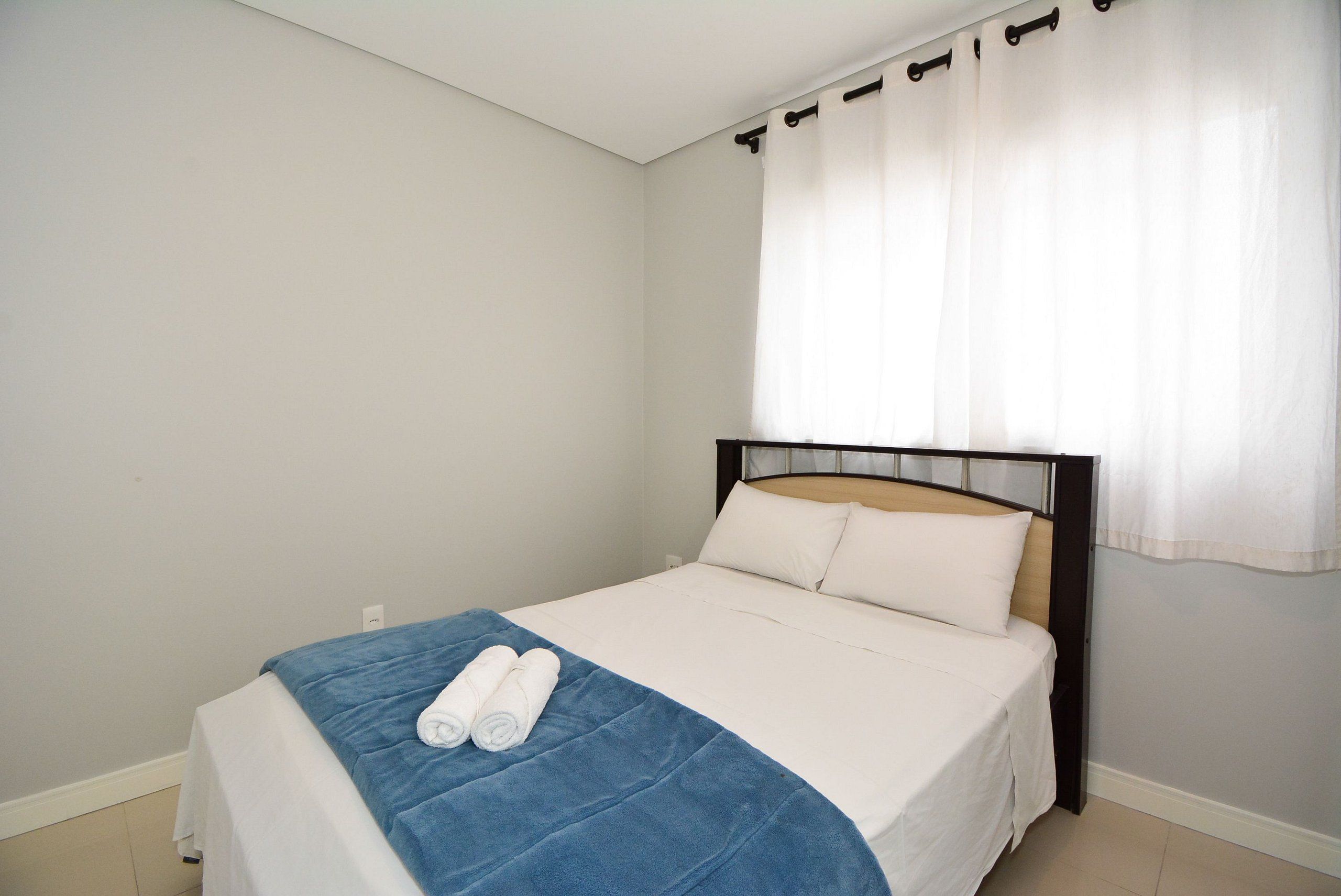 A single bed with a white mattress and blue blanket, a wooden headboard, and a window with white curtains.