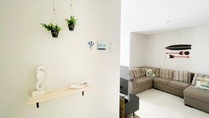 A modern living room with a sectional sofa, a wooden shelf with decorative items, and wall-mounted decorations including a seahorse, a boat, and a surfboard. - Residencial Dom Virgílio (Florianópolis)