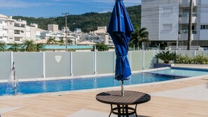 A swimming pool area with a table and umbrella, surrounded by a fence, with buildings in the background. - Residencial Dom Virgílio (Florianópolis)
