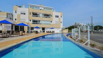 Une piscine aux carreaux bleus, entourée de chaises longues et de parasols, devant un immeuble à plusieurs étages doté de balcons.