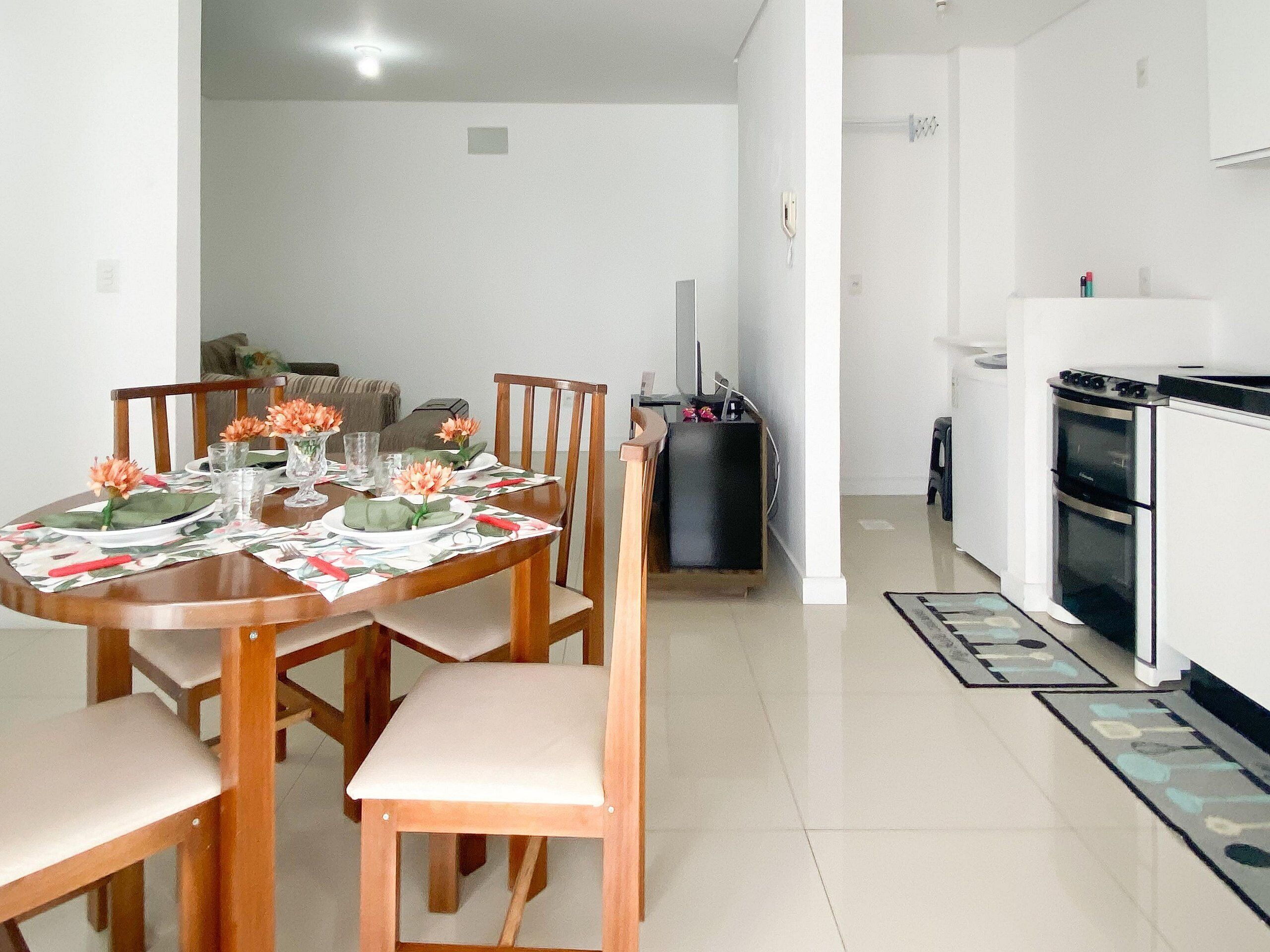 A dining area with a wooden table set for four, surrounded by chairs. A black cabinet is against the wall, and a white refrigerator is in the background.