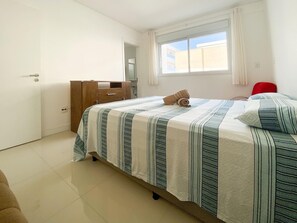A bedroom with a bed, a wooden dresser, a red chair, and a window with curtains. - Residencial Dom Virgílio (Florianópolis)