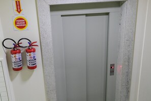 An elevator with fire extinguishers mounted on the wall and a fire exit sign above. - Residencial Dom Virgílio (Florianópolis)