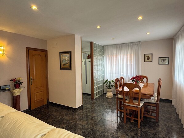 A hotel room with a bed, a dining table with chairs, a refrigerator, and a potted plant.
