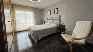 Un dormitorio con cama, una silla, una mesita de noche, una ventana con cortinas y un ventilador de techo.