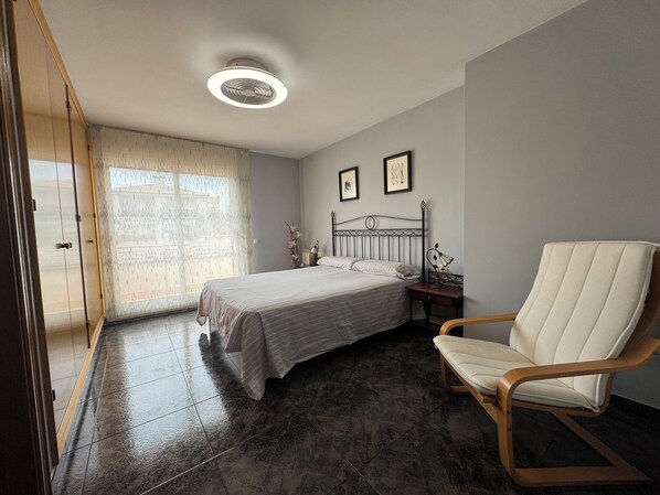 Un dormitorio con cama, una silla, una mesita de noche, una ventana con cortinas y un ventilador de techo.