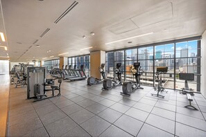 Une salle de sport spacieuse, dotée de divers équipements d’exercice, de grandes fenêtres offrant une vue sur la ville, et d’un design moderne et épuré.