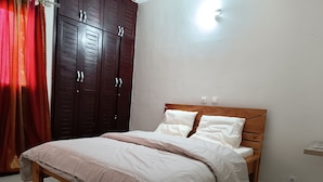A bedroom with a wooden bed, white bedding, a wardrobe, and a ceiling light. - Quiet and Secure Apartment in Angré 9e Tranche, Abidjan, A Haven of Peace. (Abidjan Cocody Angre 9ieme Tr.)