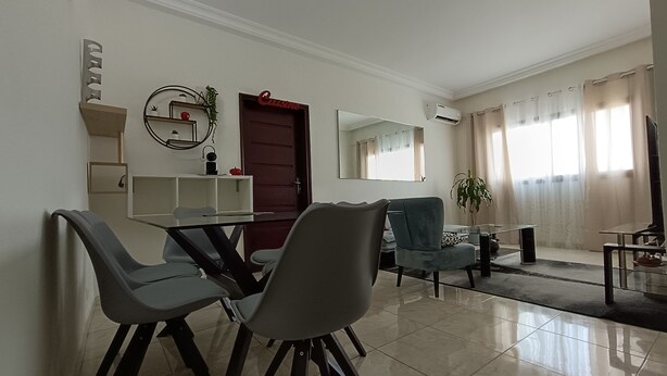 A modern living room with a dining area, featuring a glass table, grey chairs, a blue armchair, and a large mirror. - Quiet and Secure Apartment in Angré 9e Tranche, Abidjan, A Haven of Peace. (Abidjan Cocody Angre 9ieme Tr.)