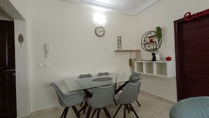 A dining area with a glass table and four chairs, a wall clock, a phone, and a shelf with decorative items. - Quiet and Secure Apartment in Angré 9e Tranche, Abidjan, A Haven of Peace. (Abidjan Cocody Angre 9ieme Tr.)