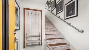 Una escalera de madera con barandilla metálica que lleva a un rellano con una puerta blanca y un estante con cuadros enmarcados.