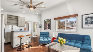 Una sala moderna con un sofá azul, una mesa de centro con flores, y una zona de cocina con un taburete de bar y un ventilador de techo.