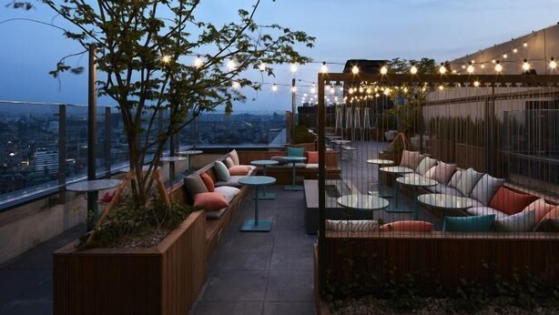 An outdoor seating area with cushioned benches and round tables, surrounded by plants and string lights.