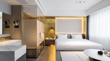A modern hotel room with a large bed, a separate bathroom, a round dining table, and a large window.