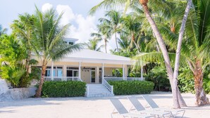 A beachfront property with a covered porch, palm trees, and lounge chairs on the sand. - Island Palm Estate in Islamorada (Islamorada)