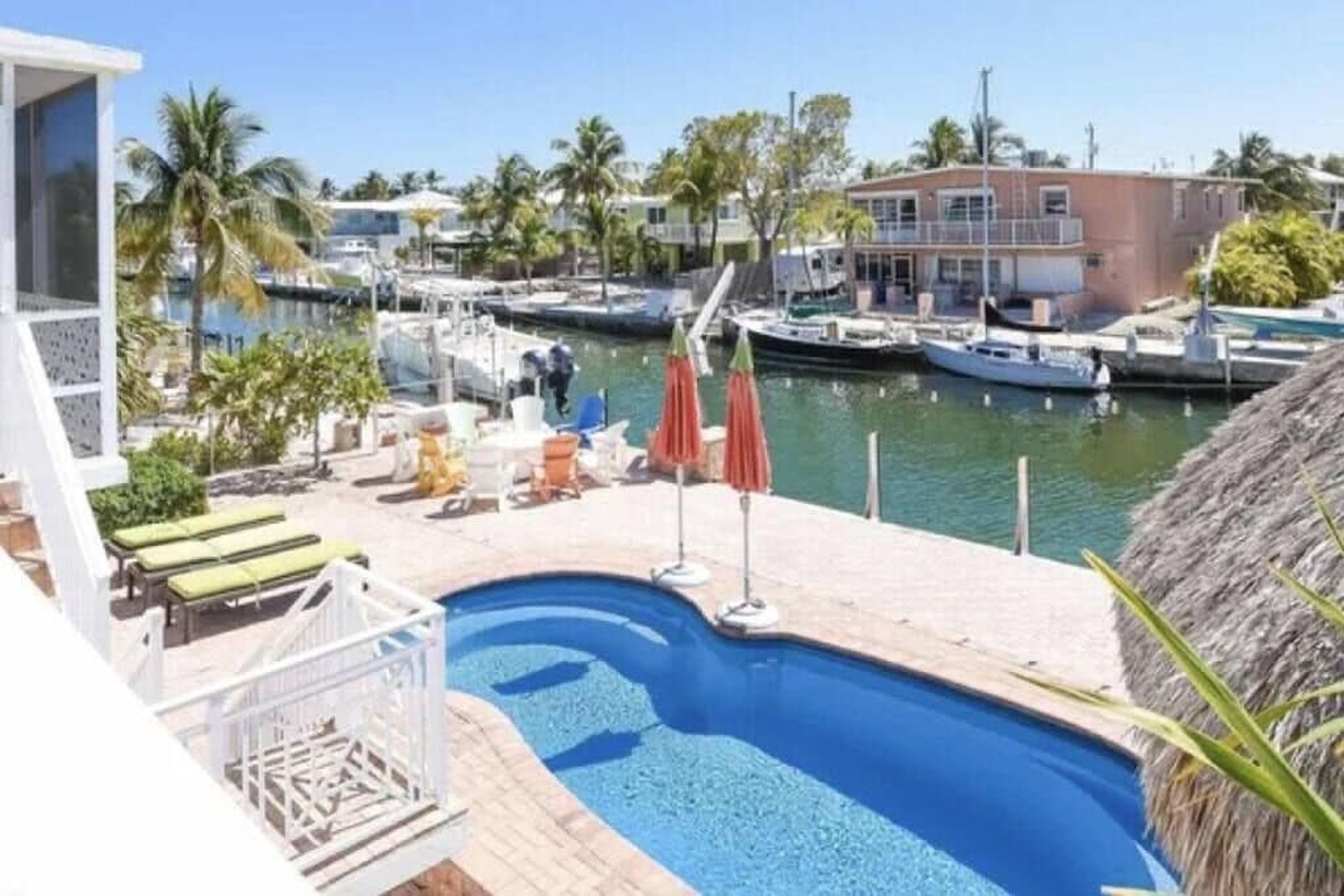 Een zwembad met ligbedden en parasols, omgeven door een jachthaven met boten en palmbomen.