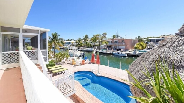 A pool area with a view of the marina, a thatched roof structure, and a building with a balcony. - Holly s Hideout in Islamorada (Tavernier)