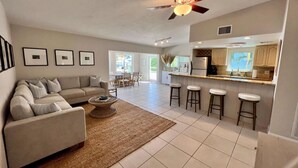 A modern living room with a sofa, coffee table, and a ceiling fan. There is a kitchen area with bar stools and a dining area visible in the background. - Boaters Retreat in Islamorada (Islamorada)