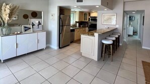 A modern kitchen with white cabinetry, a central island with bar stools, and a visible stainless steel refrigerator. - Boaters Retreat in Islamorada (Islamorada)