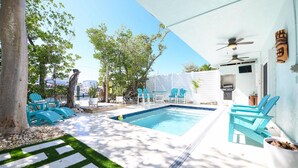 Un área de piscina con una piscina azul cristalina, rodeada de muros blancos y una cerca blanca. Varias sillas azules y una mesa se encuentran bajo un dosel blanco.