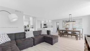 A modern living room with a sectional sofa, a dining area with a wooden table and benches, and a kitchen in the background.
