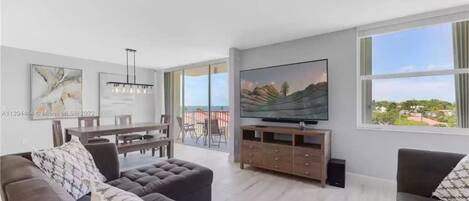 A spacious living room with a large TV, comfortable seating, and a balcony with a scenic view.