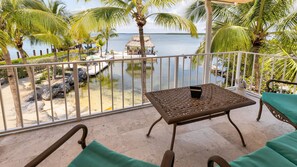A balcony with a table and chairs, overlooking a beach with palm trees and a thatched-roof hut.