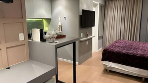Una habitación de hotel moderna con cama, escritorio, televisor de pantalla plana, kitchenette pequeña y un ventanal con cortinas.
