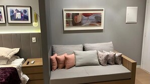 Un dormitorio moderno con un sofá gris, una mesita de noche con lámpara y cuadros en la pared.