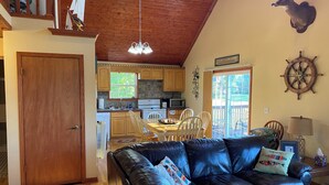 A cozy living room with a leather sofa, wooden dining table, and a kitchen in the background. - Charming 3-BR house w/golf, pool, WiFi, AC at pet friendly Middle Bass Island (Middle Bass)