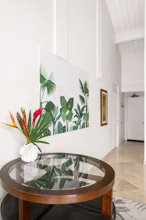 A room with a glass-top table, a white flower vase, a tropical-themed wall art, and a framed picture.