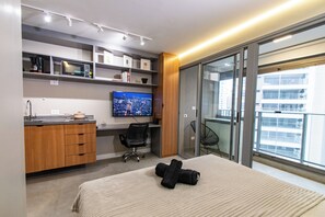A modern hotel room with a bed, a desk with a computer, a TV, and a balcony with a city view.