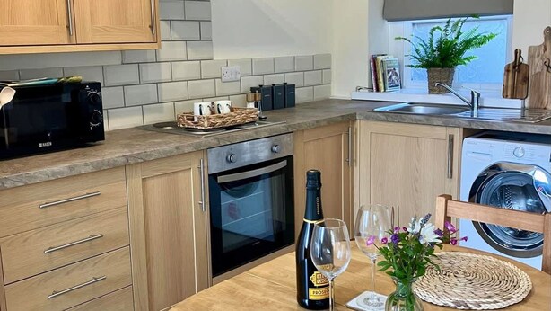 A modern kitchen with wooden cabinets, a built-in oven, a microwave, and a dining area with a round table and woven placemats. - Farmhouse Annex in Saint Ishmael s (Haverfordwest)