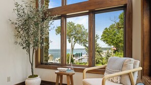 Una habitación con un ventanal grande que ofrece vistas a un muelle y árboles, una planta en maceta, una mesita con un cuenco y un objeto decorativo, y un sillón acolchado.