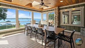 Un patio techado con una mesa y sillas de madera, amplias ventanas que ofrecen vistas al lago y un ventilador de techo.