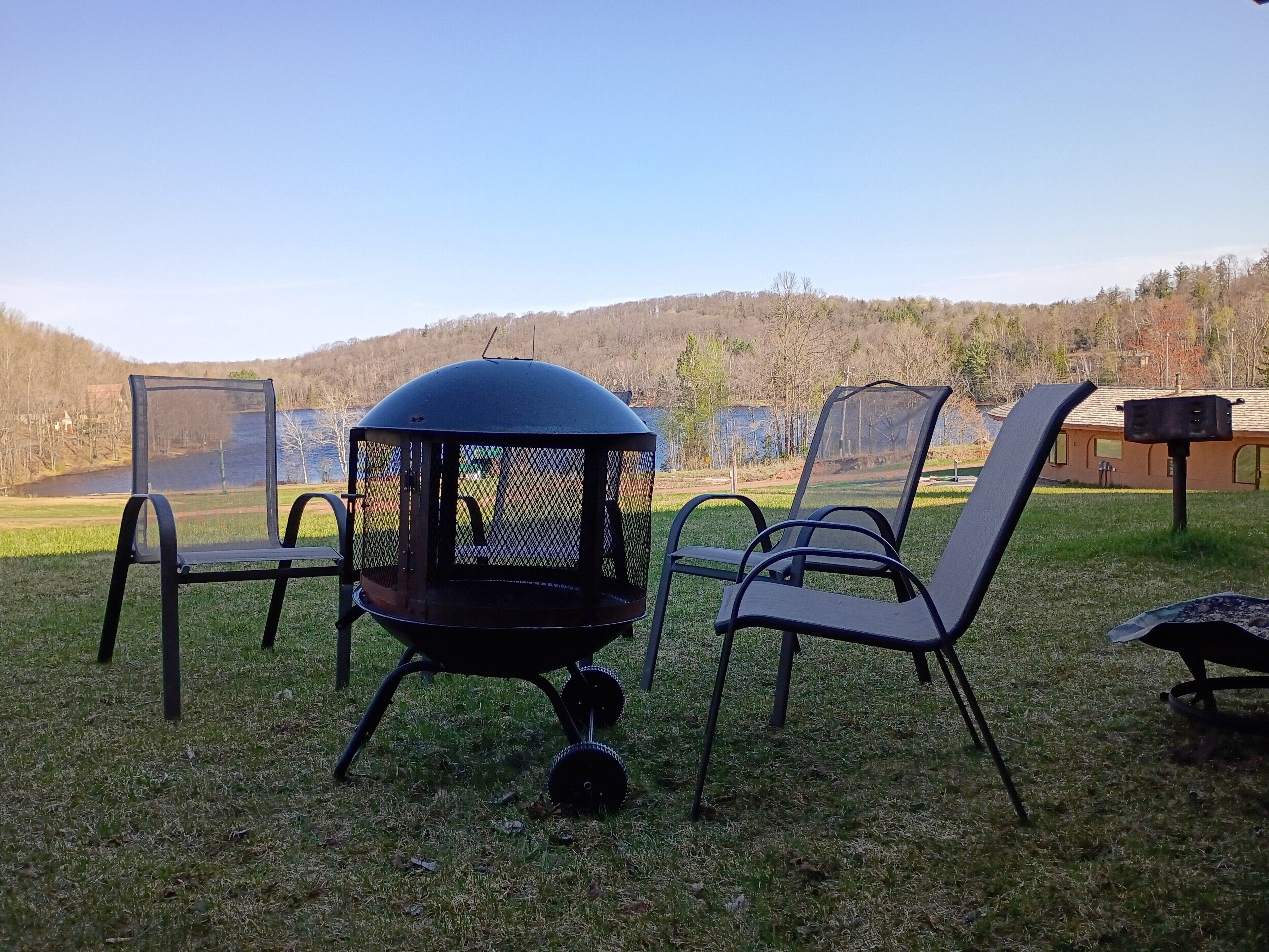 A grill with a mesh cover and a chimney, surrounded by metal chairs on a grassy area with a view of a lake and hills in the background.