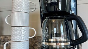 A coffee maker with a digital display and a stack of white mugs with textured designs. - Camp Street Haven: 2-bedroom peaceful getaway (Ely)