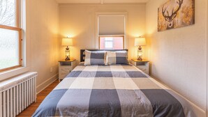 A bedroom with a large bed, two bedside tables with lamps, a window with blinds, and a deer painting on the wall. - Camp Street Haven: 2-bedroom peaceful getaway (Ely)