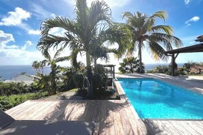 A wooden deck with a pool, palm trees, and a view of the ocean.