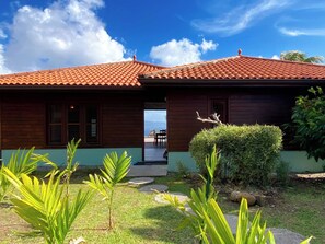 A wooden house with a red-tiled roof, a small garden with palm plants, and a view of the ocean.