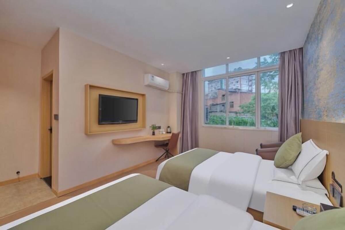 A hotel room with two beds, a TV, a desk, and a window with a view of buildings and trees.