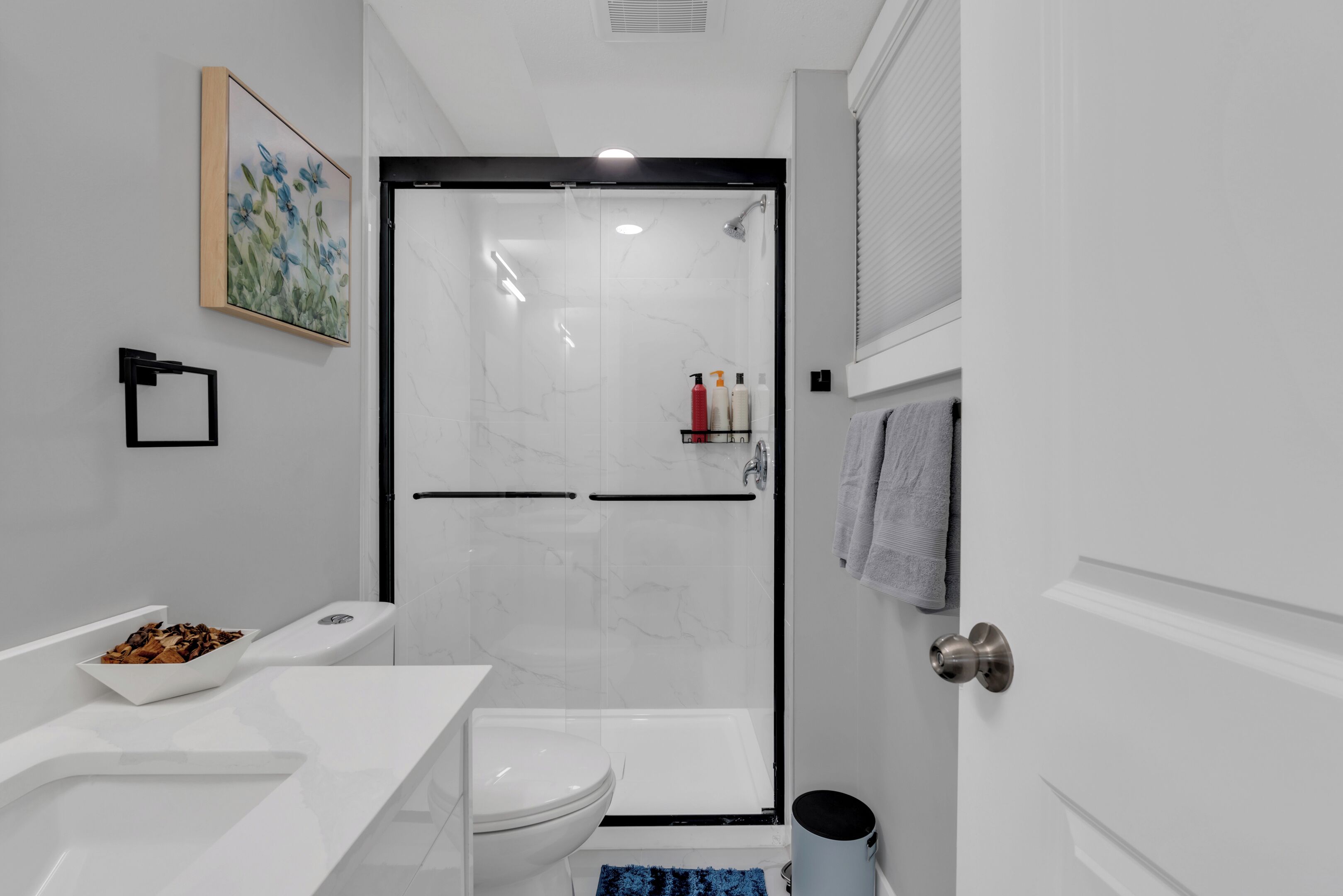 A modern bathroom with a white sink, toilet, and shower area. There is a framed picture on the wall and a towel rack with towels.