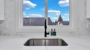 Una cocina con ventana, un lavamanos de acero inoxidable y dos botellas de detergente.