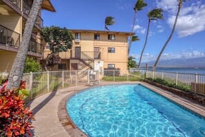 A swimming pool surrounded by a white fence, with a building in the background and palm trees. - Suite Maui Paradise Condo (Wailuku)