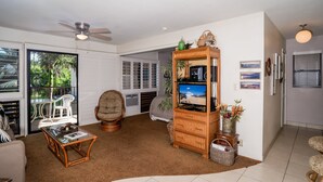A spacious living room with a ceiling fan, a large TV, wicker furniture, and a view of the outdoors through glass doors. - Suite Maui Paradise Condo (Wailuku)