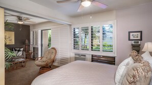 A bedroom with a large bed, a ceiling fan, a window with blinds, a chair, and a nightstand with a lamp. - Suite Maui Paradise Condo (Wailuku)