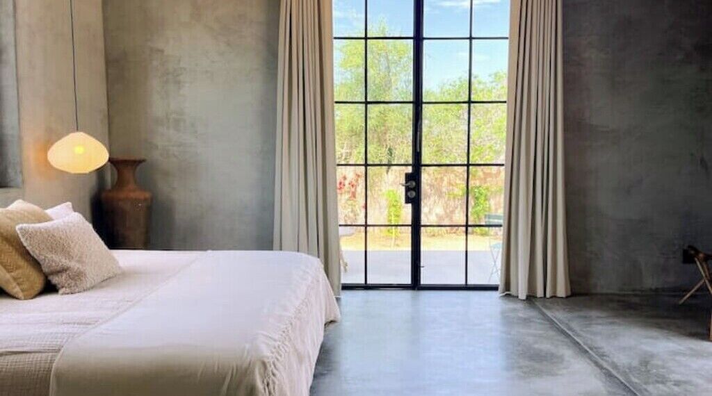 A bedroom with a large bed, a nightstand, a lamp, and a view through a glass door to the outdoors.