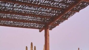 Un área de comedor al aire libre con mesas y bancos de madera, una piscina y cactus al fondo.